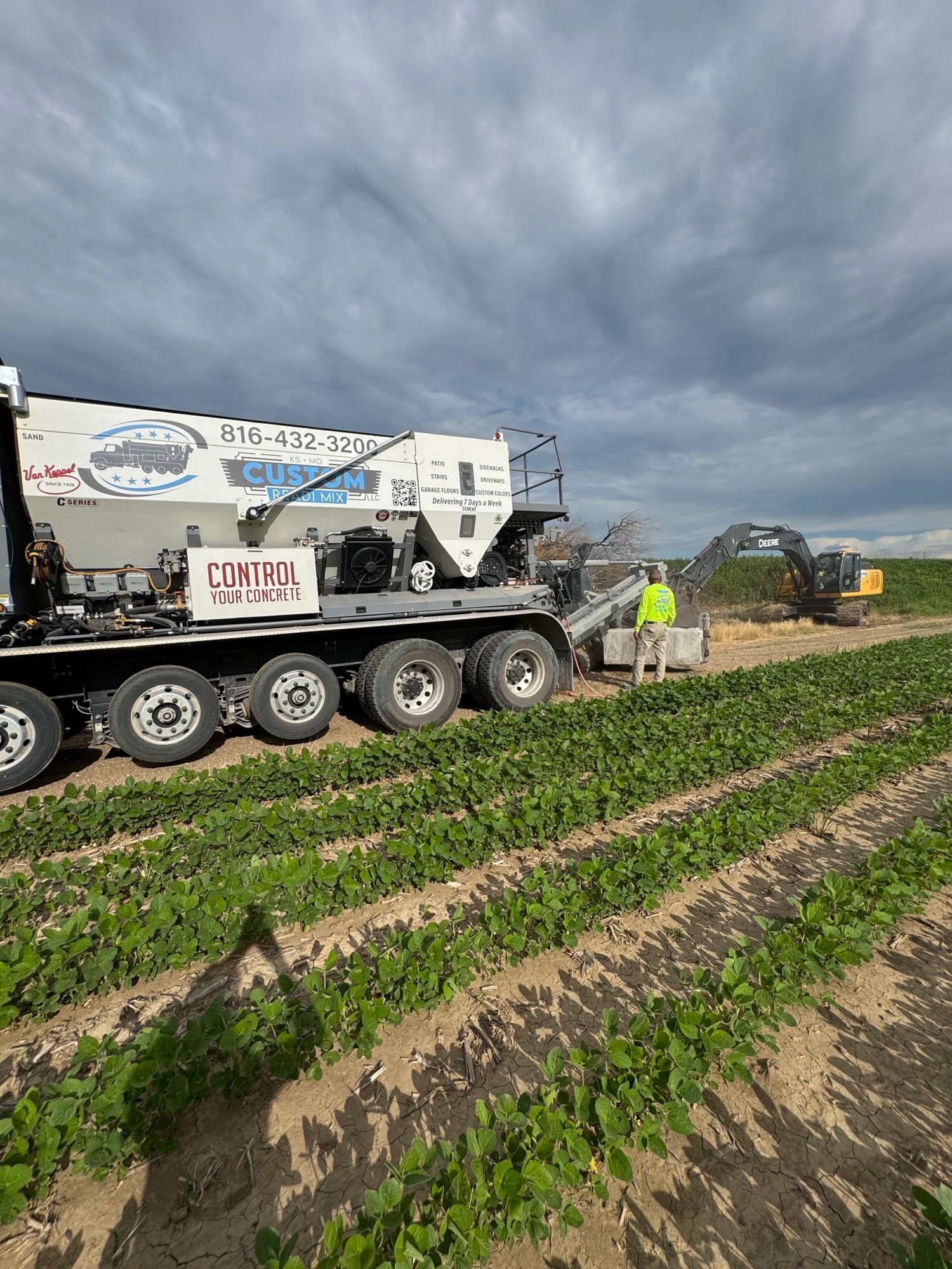 A large white truck dispensing material into a field with rows of green plants, under a cloudy sky.