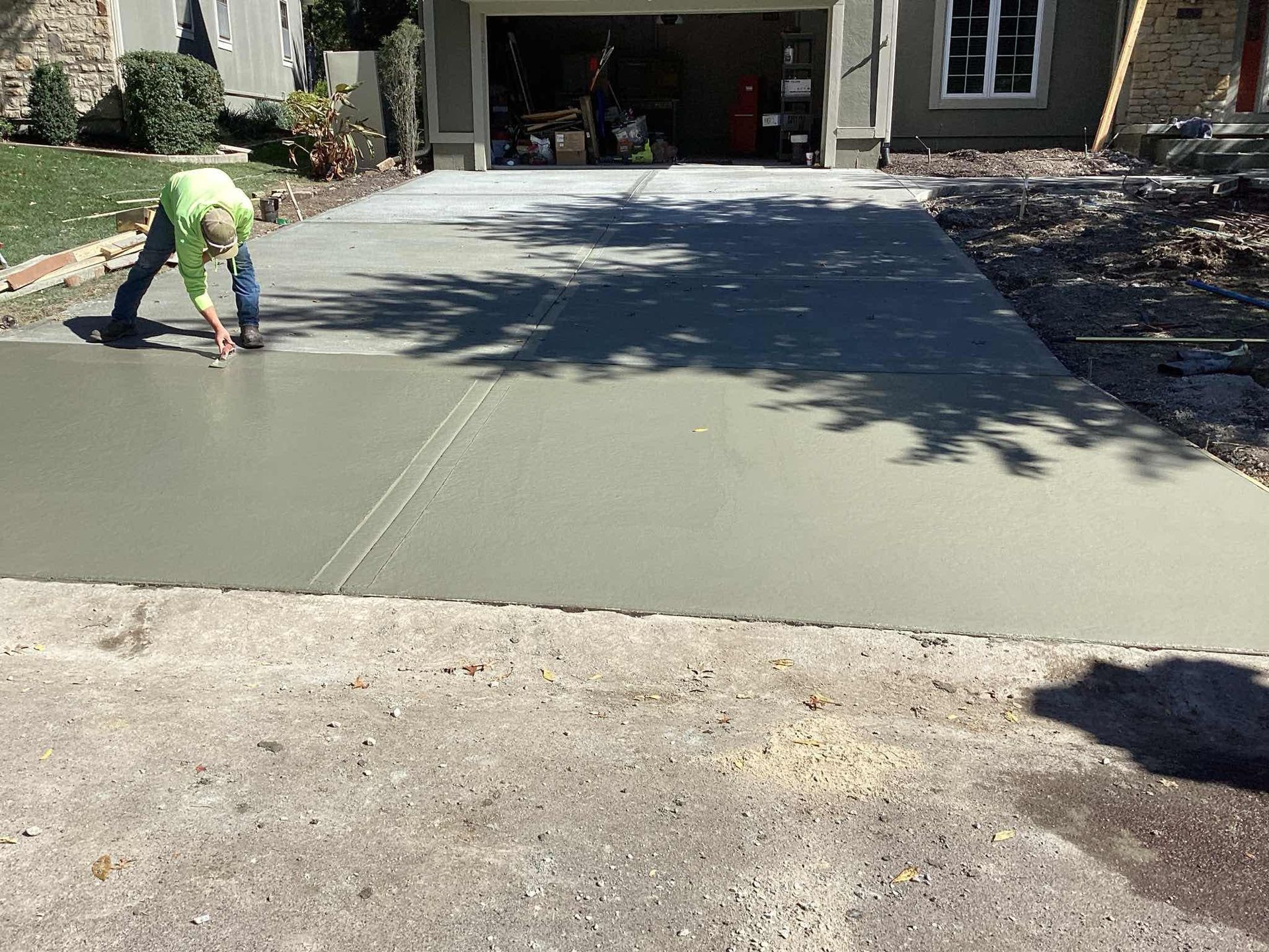 Person in green shirt smoothing wet concrete driveway near a garage.
