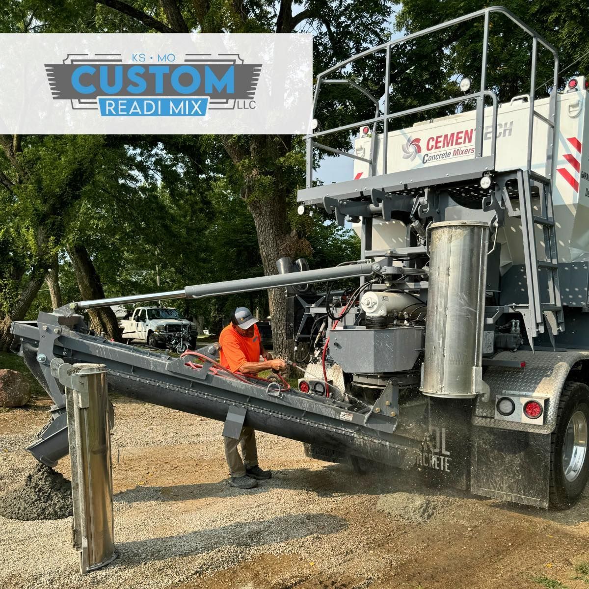 A person in an orange shirt operates a concrete conveyor truck pouring material onto a gravel surface.