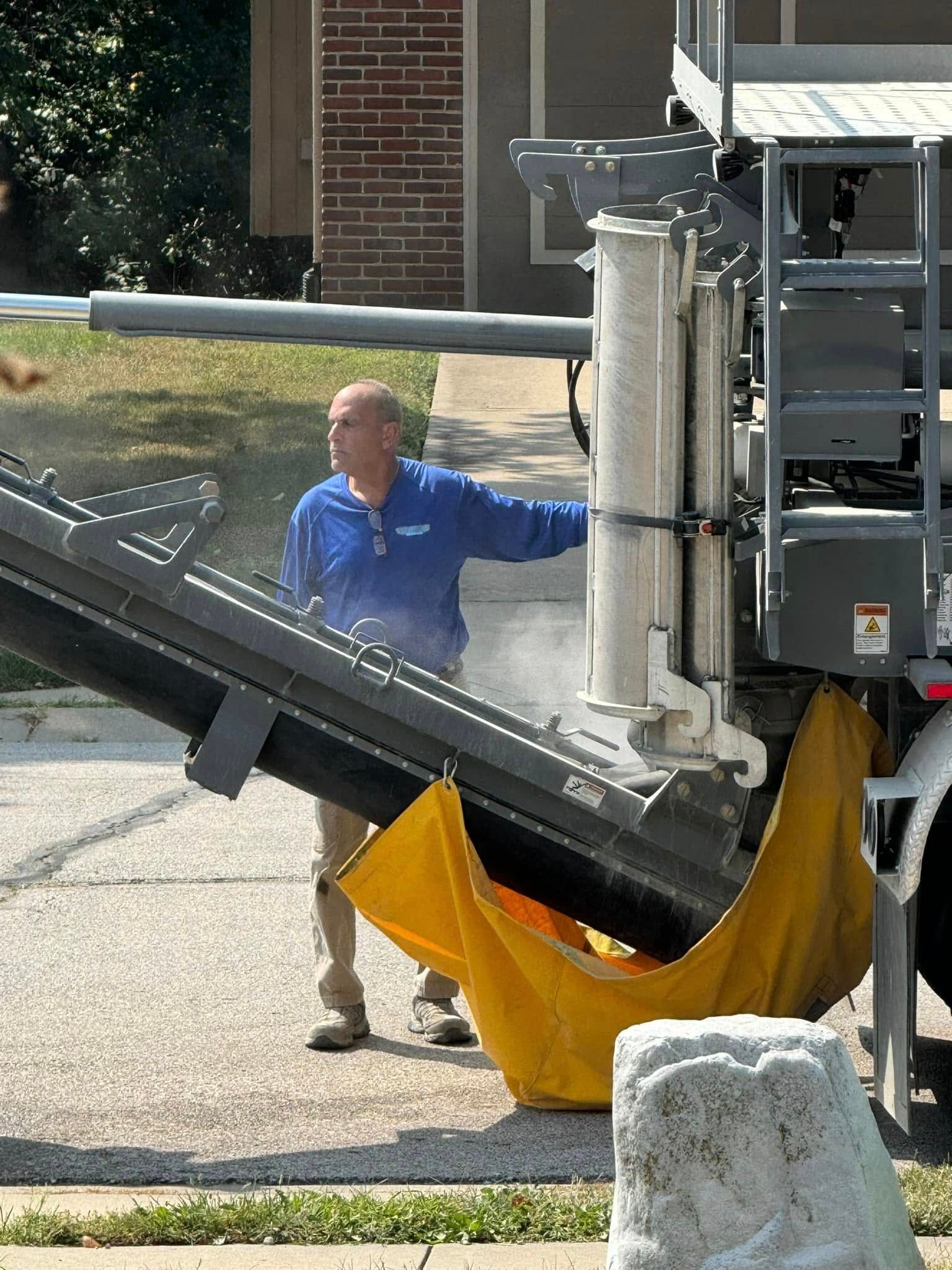 A person in a blue shirt stands next to industrial construction equipment with a large yellow dust shroud on a paved road.