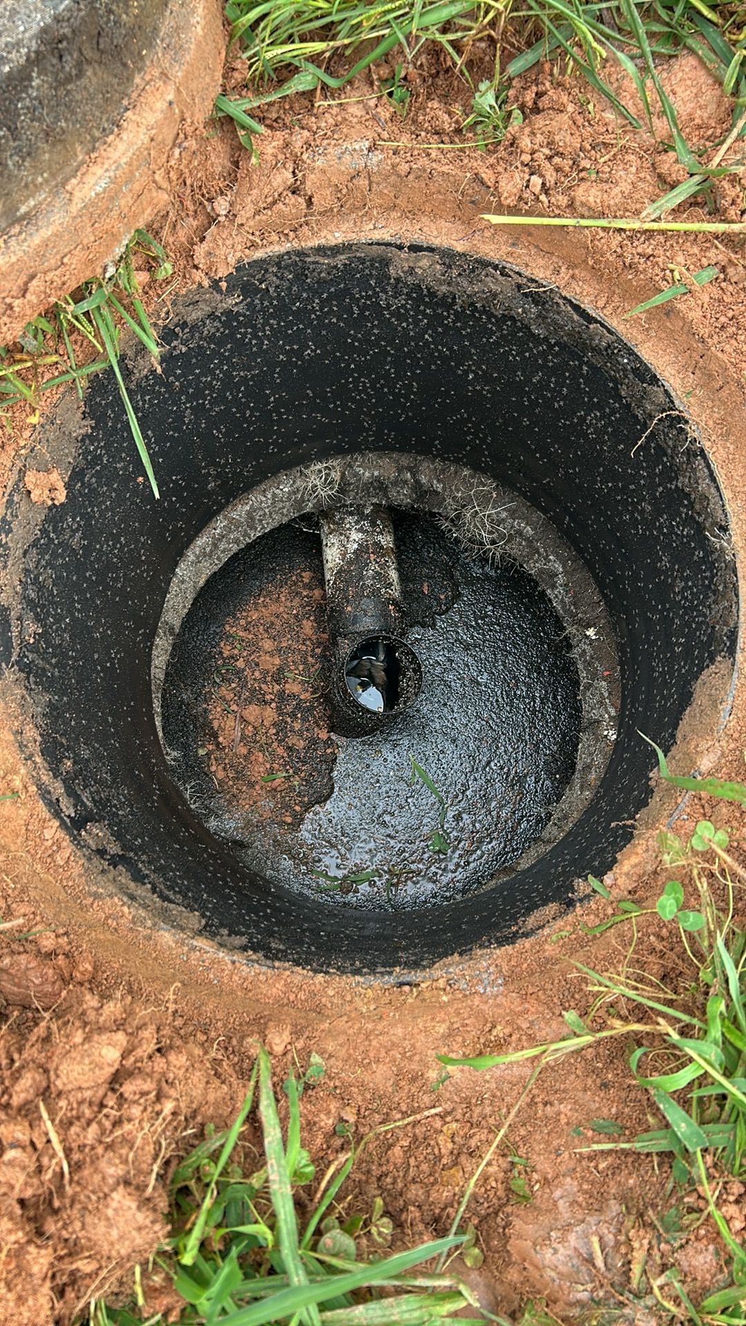 Open sewer access point, showing a dark interior with standing water and a pipe. The surrounding ground is dirt and grass.