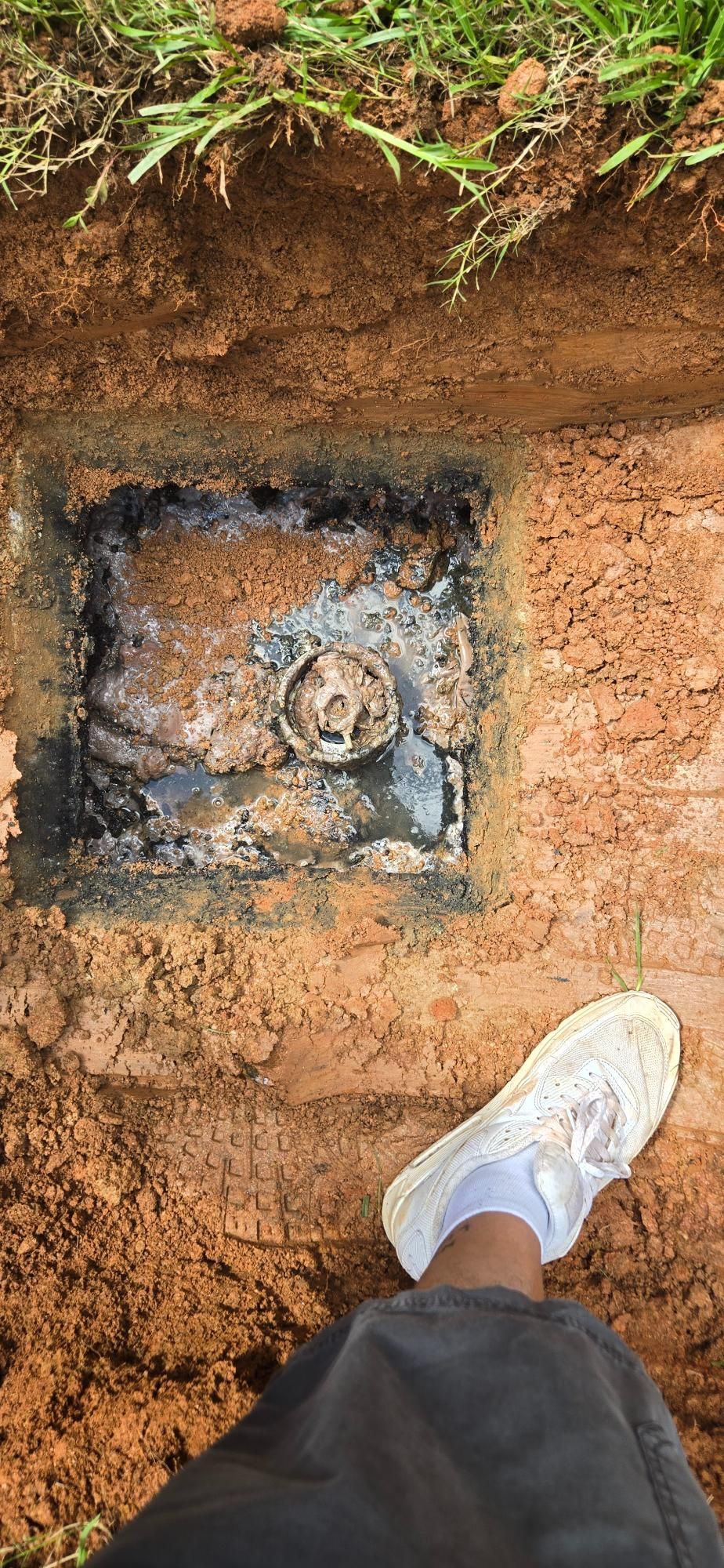 A foot wearing a white sneaker beside a square opening in the ground, revealing a pipe surrounded by dirt.