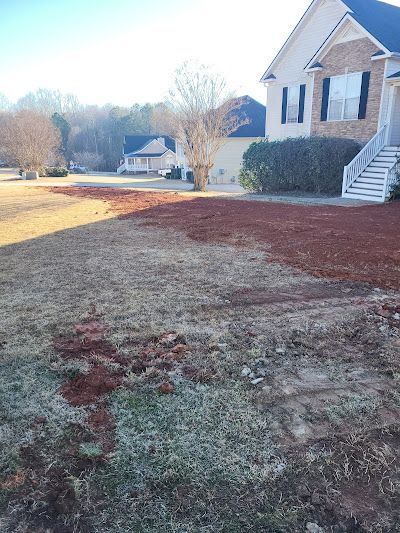 A front yard with patchy brown grass and sections covered in reddish-brown mulch, in front of a two-story house.
