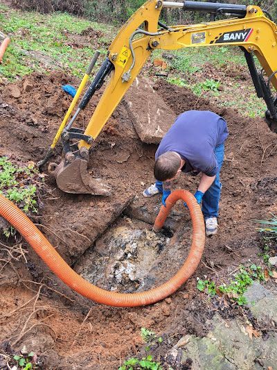 A person in blue leans over a hole in the ground next to a yellow excavator, with an orange hose inserted into the hole.