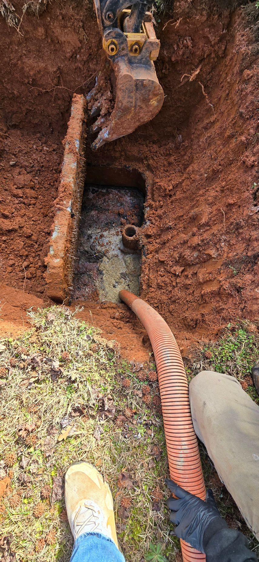 An excavator digs near a brick structure, with an orange corrugated hose extending from the hole. 