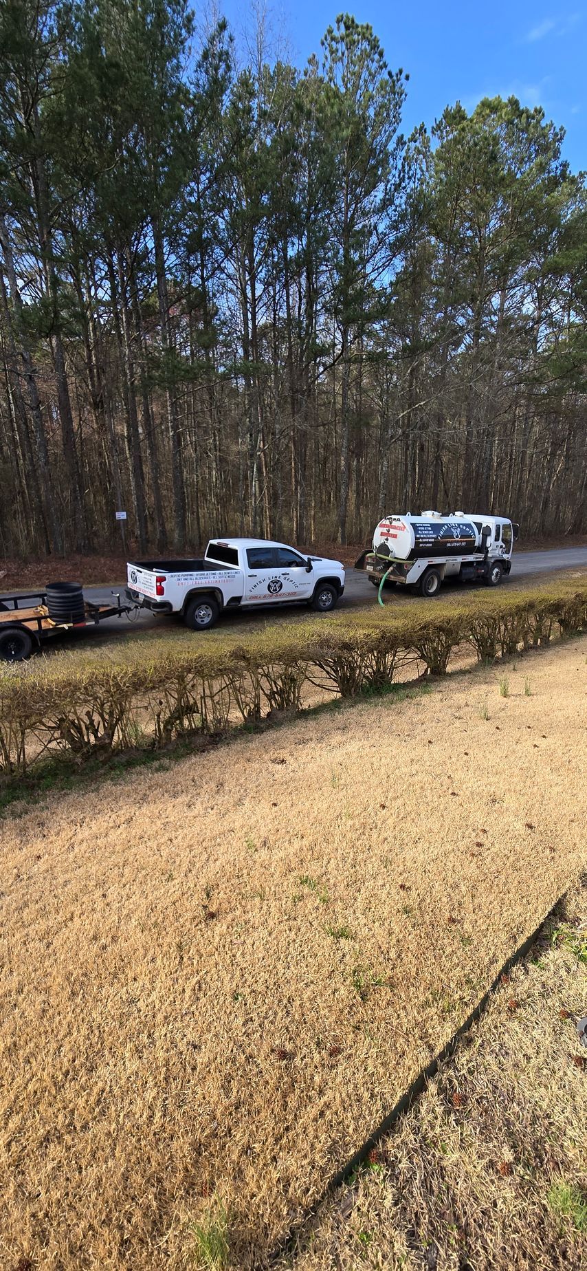 A white pickup truck towing a trailer and a camper drives on a road next to a brown grass field.