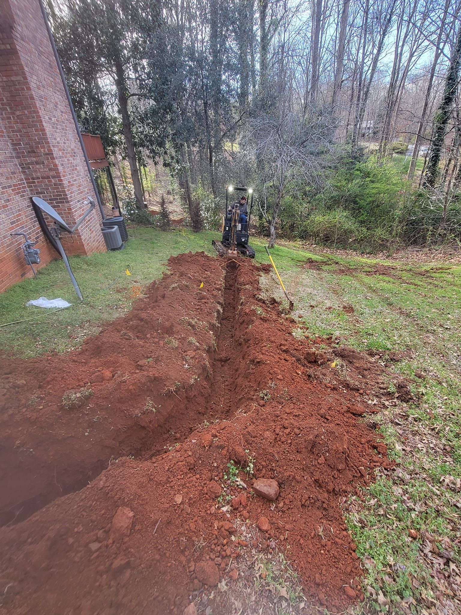 A trench being dug by a small backhoe in a grassy yard near a brick building and trees; red soil is piled alongside.