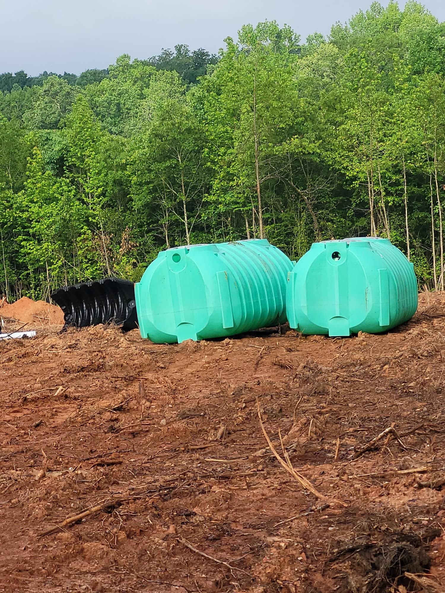 Two green septic tanks on a dirt lot with trees in the background. One black tank is also visible.