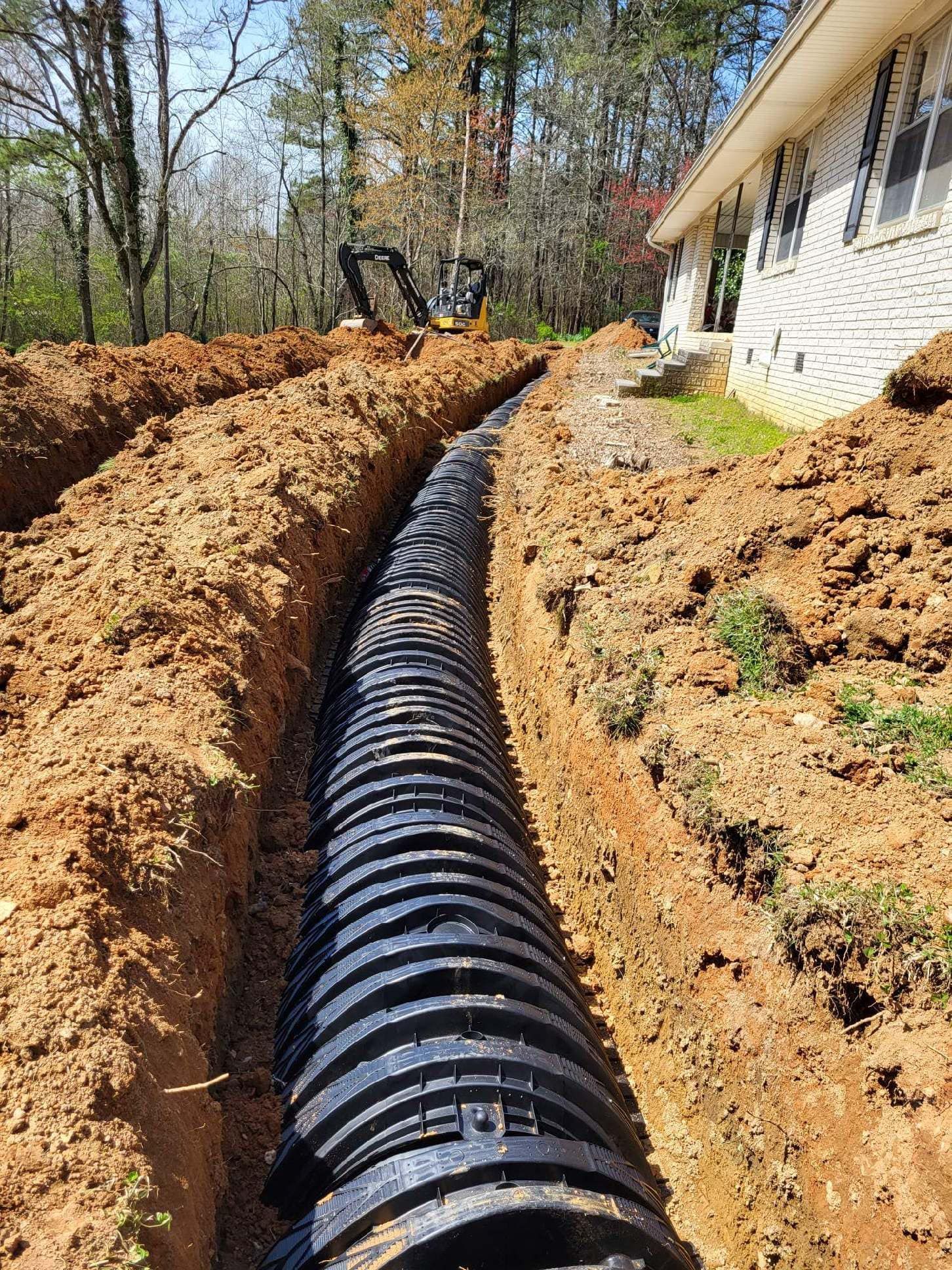 A black corrugated pipe installed in a trench along the side of a white brick house. Dirt is piled on either side.