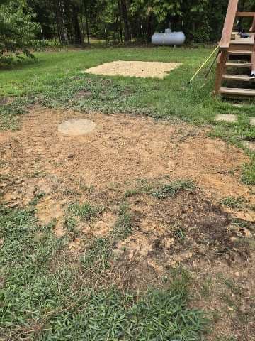 A cleared dirt area surrounded by grass, a small gravel patch in the background, and a propane tank; outdoors.