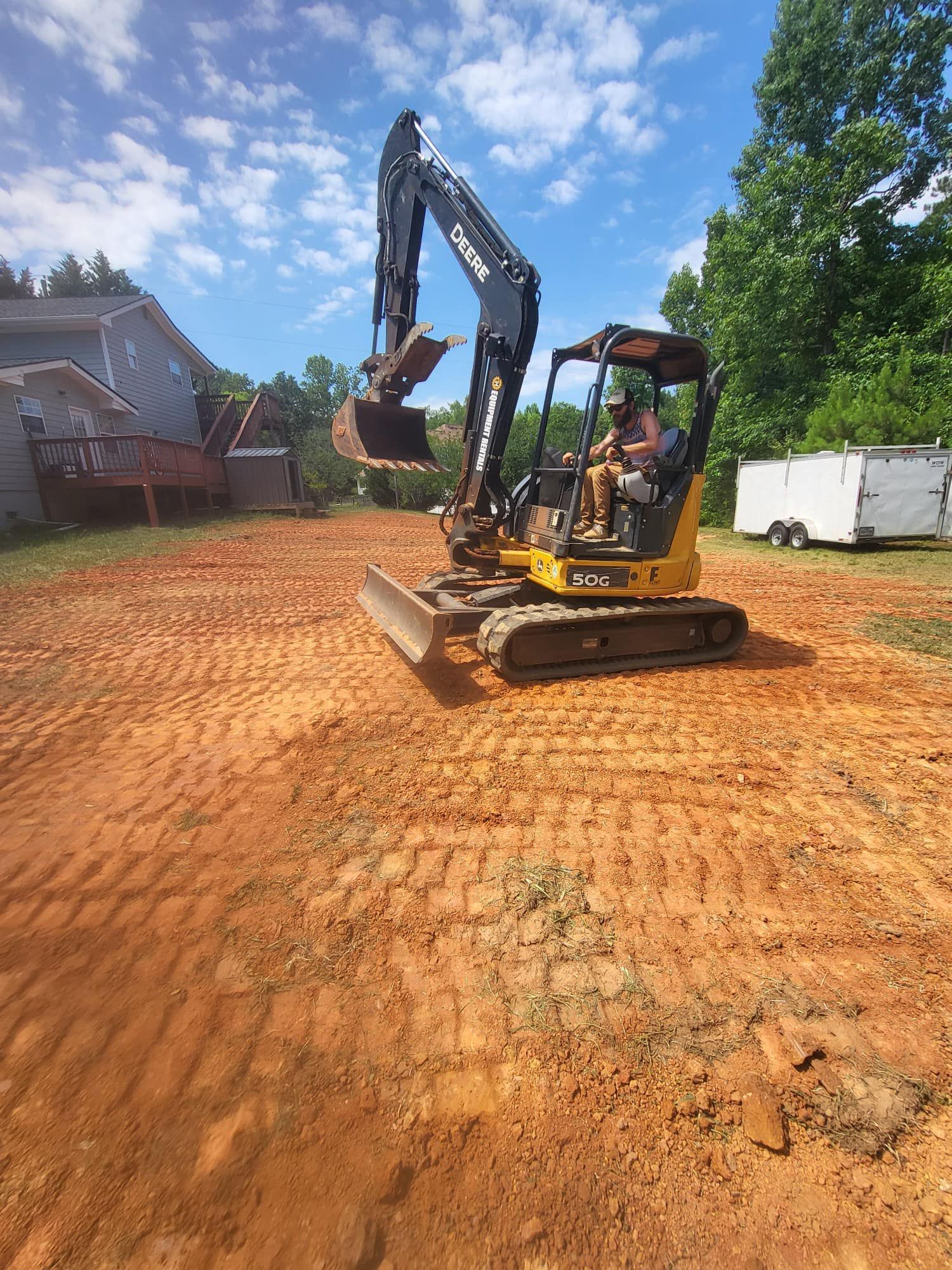 A small excavator on a cleared dirt lot, driven by a person, with a house and trees in the background.