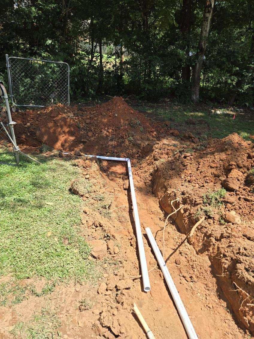 A trench dug in a grassy yard with buried pipes for a construction project near a fence and trees.