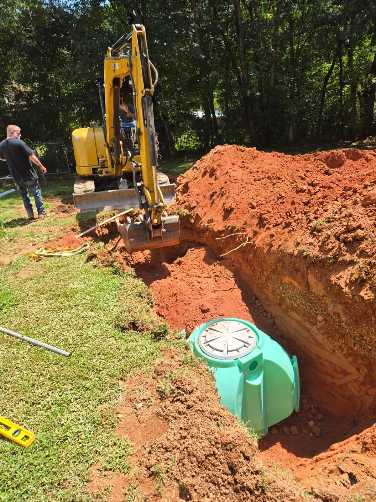 An excavator installing a green septic tank in a red clay trench, with a worker observing on the grass.