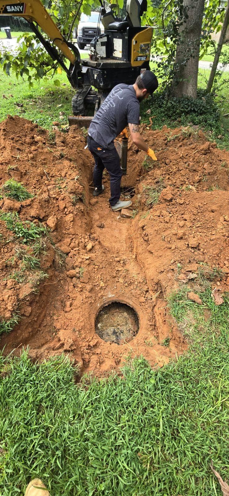 A person digging near an open circular access point in the ground. A yellow mini-excavator is behind them.