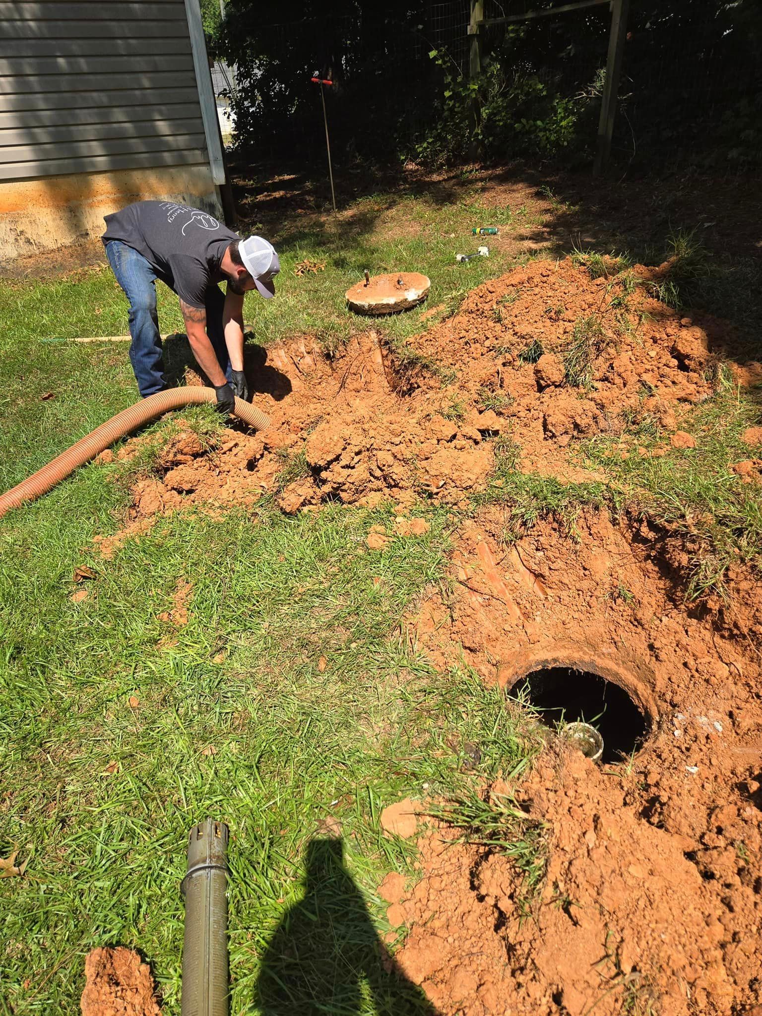 Man pumping out a septic tank in a yard; he is wearing jeans and a hat, dirt is piled around the open tank.