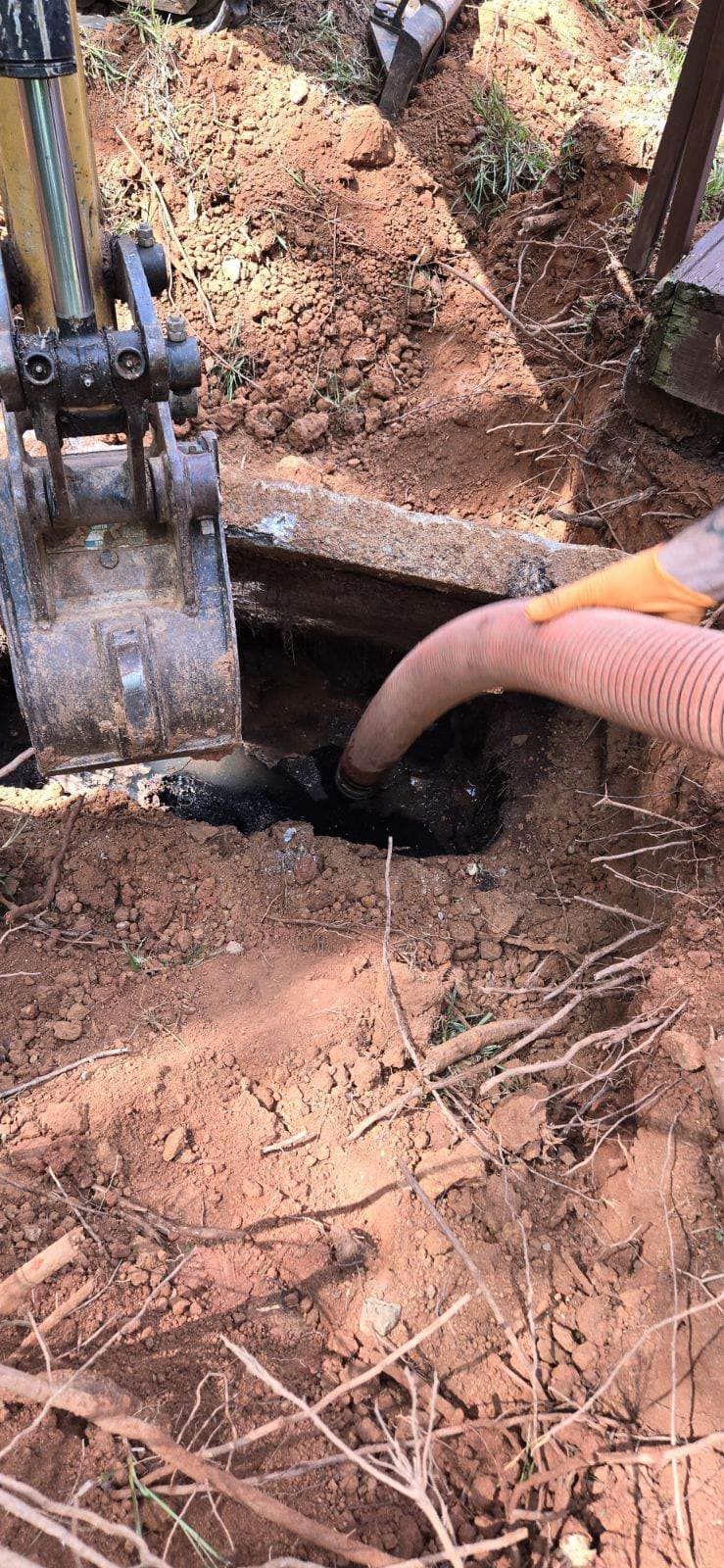 An excavator and a hose are positioned over a hole in the ground, likely extracting waste. The soil is reddish-brown.