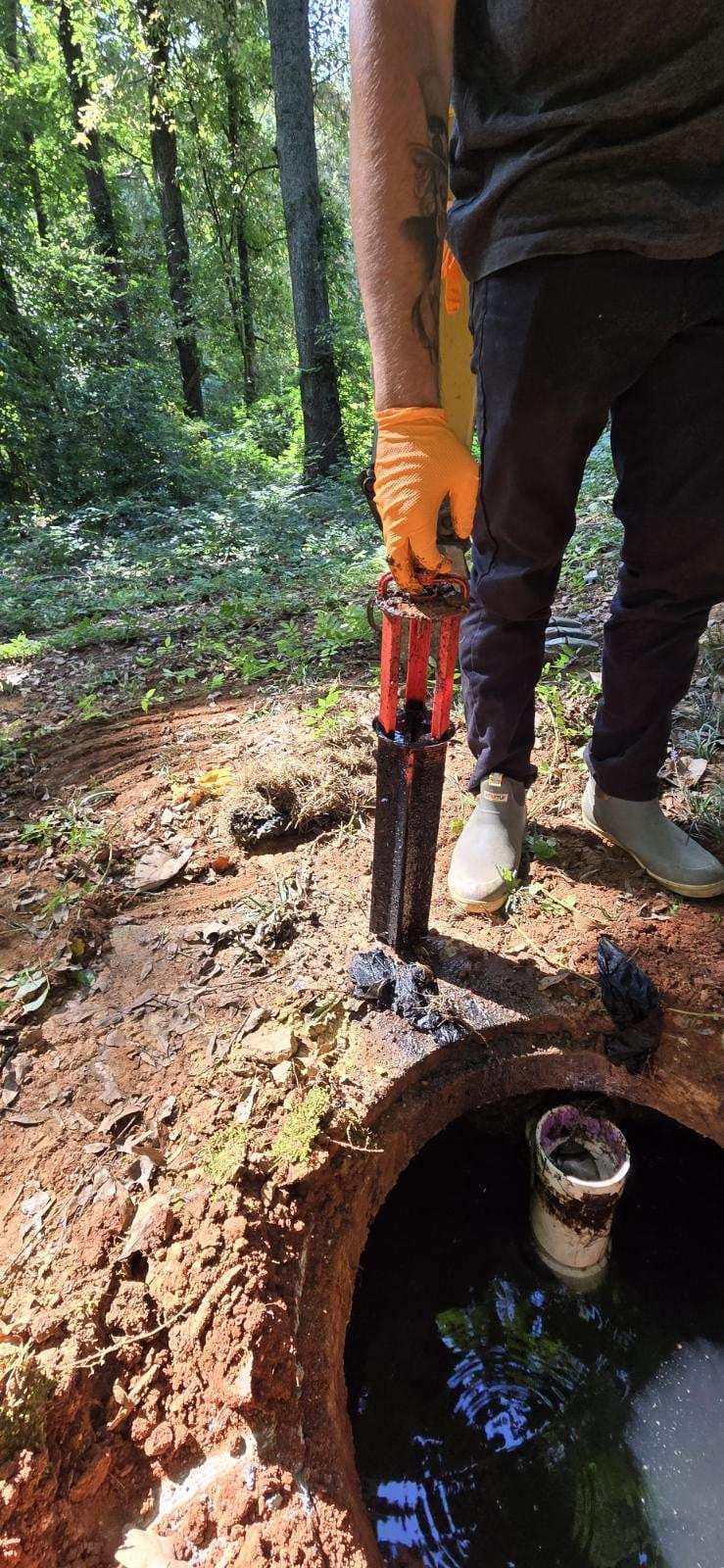 A person wearing gloves inspects a septic tank in a wooded area. They are holding a tool to measure tank contents.