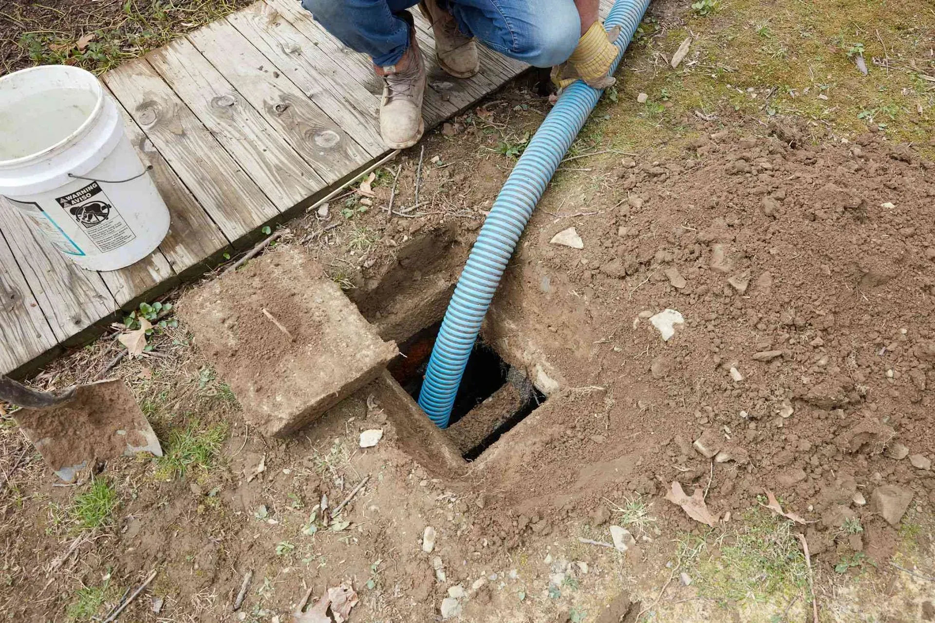 Person pumping waste from a septic tank with a blue hose in a grassy area. A bucket sits nearby.