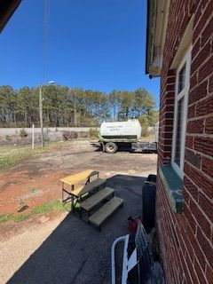 A water tanker trailer parked near a red brick building on a sunny day. A wooden bench and steps are in the foreground.