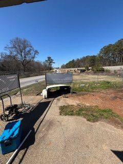 A curb ramp with a trailer on it. Gray asphalt transitions to dry grass. A fence and roadway are in the background.