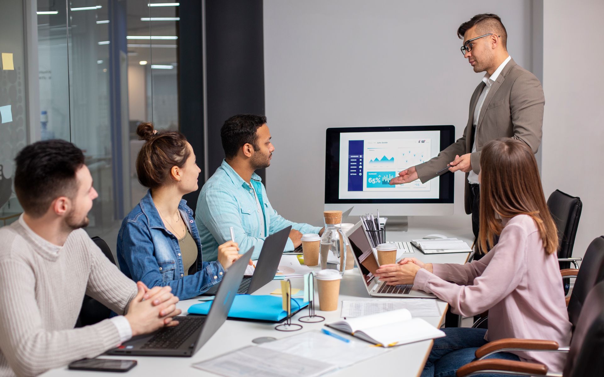 People in a business meeting, looking at a monitor. A man stands, gesturing to the screen.