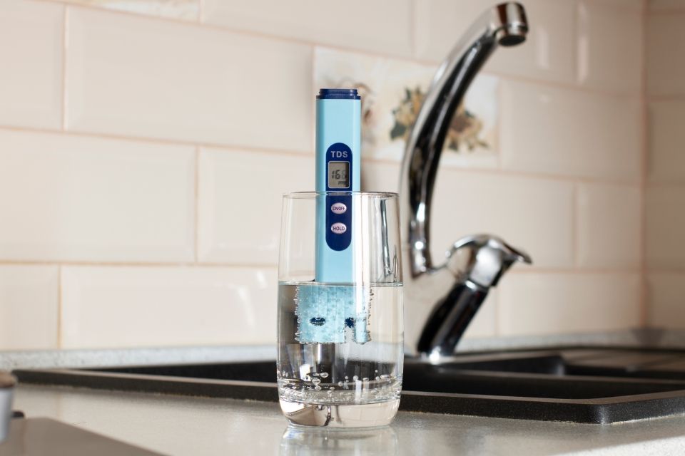 A water quality tester in a glass of water, next to a kitchen faucet, in front of tile.