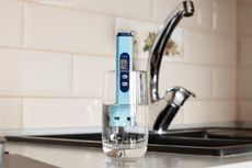 A water quality tester in a glass of water, next to a kitchen faucet, in front of tile.