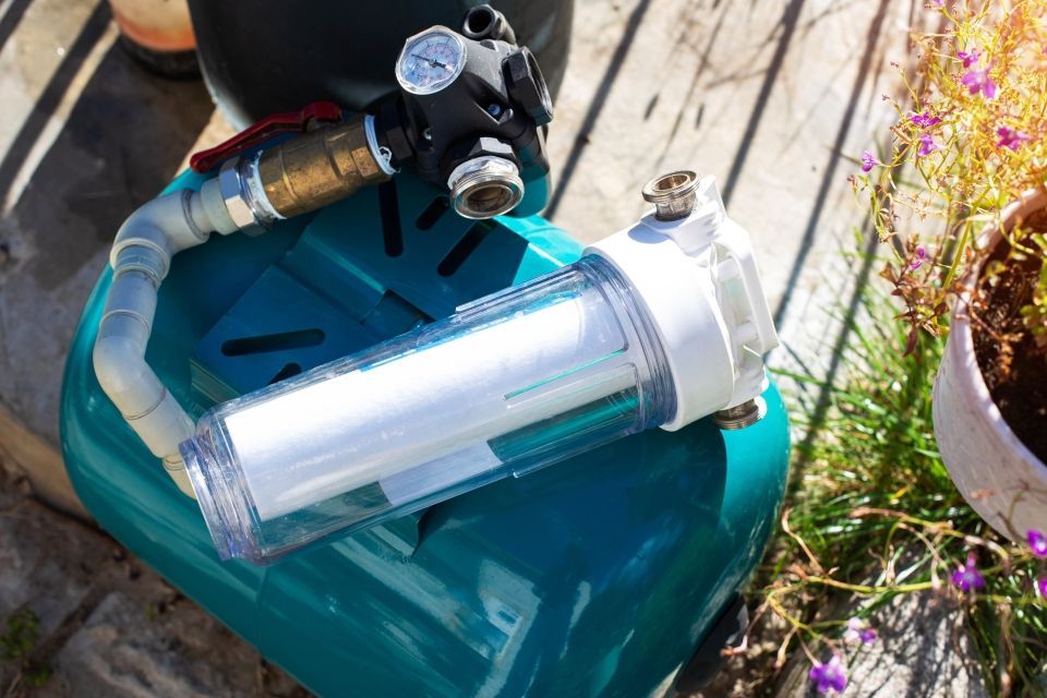 Water filtration system components on a teal container: filter housing, gauge, tubing, and valve outdoors.