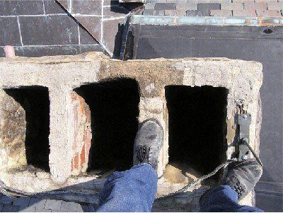 Foot on a chimney; close-up view from above.  Person in jeans & boots, roof, dark rectangular openings.