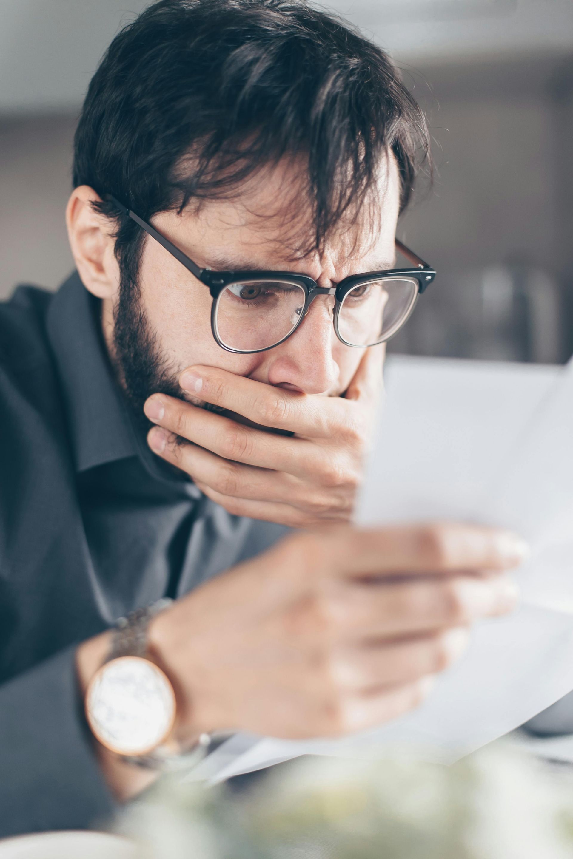 A man wearing glasses is covering his mouth while reading a letter.