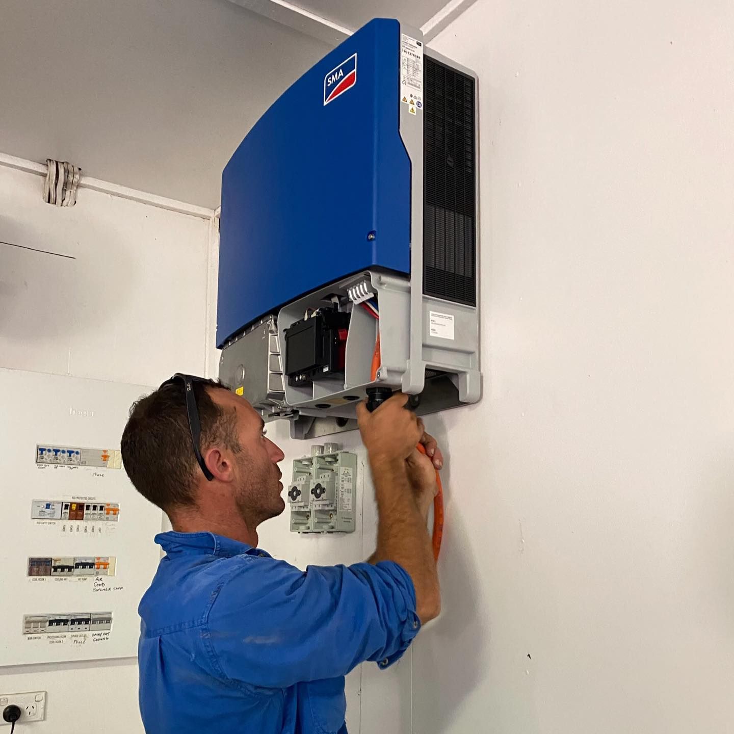 Man in Blue Jumpsuit Installing a Blue and Black Inverter — Lahtinen Electrical & Refrigeration in Ingham, QLD