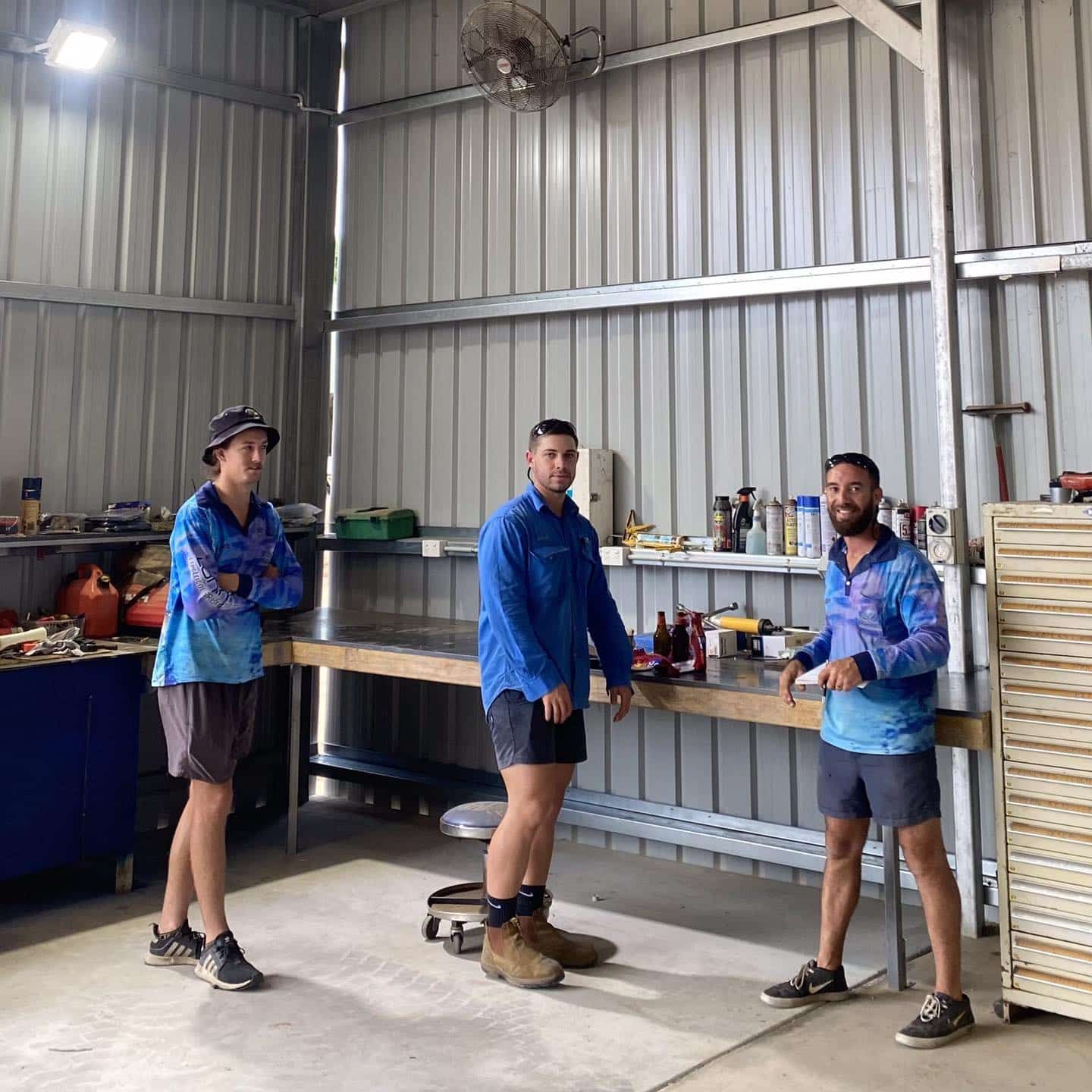 Three Men in Blue Shirts and Shorts Stand in a Workshop With Tools — Lahtinen Electrical & Refrigeration in Ingham, QLD