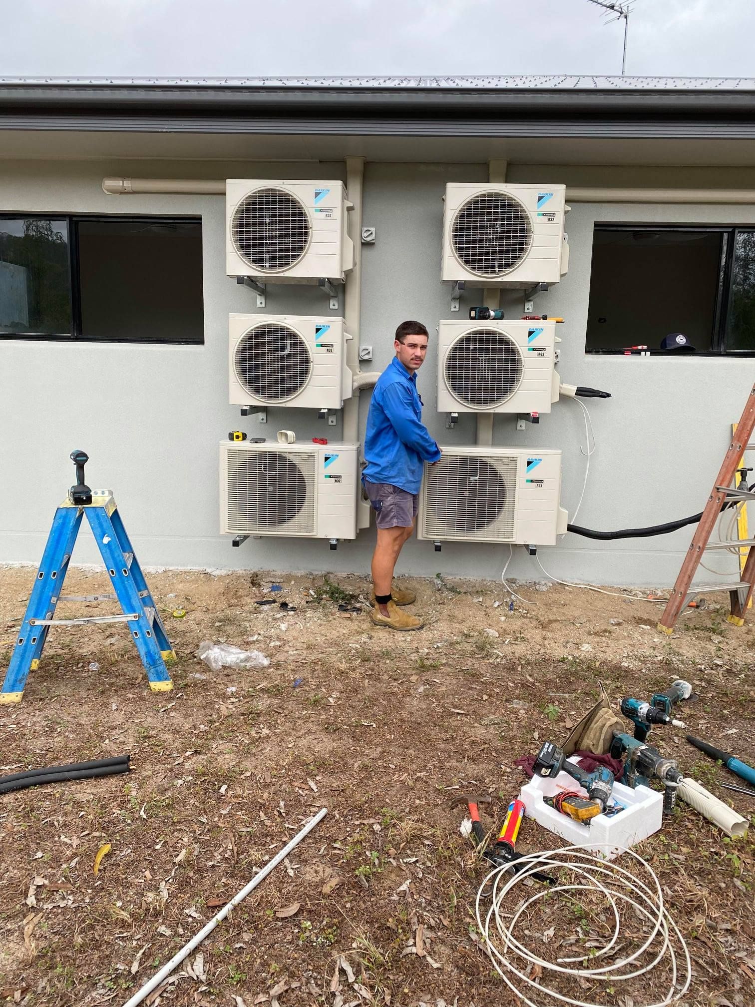 A person installing multiple air conditioning units on an exterior wall. Tools and ladder are nearby.— Lahtinen Electrical & Refrigeration in Ingham, QLD
