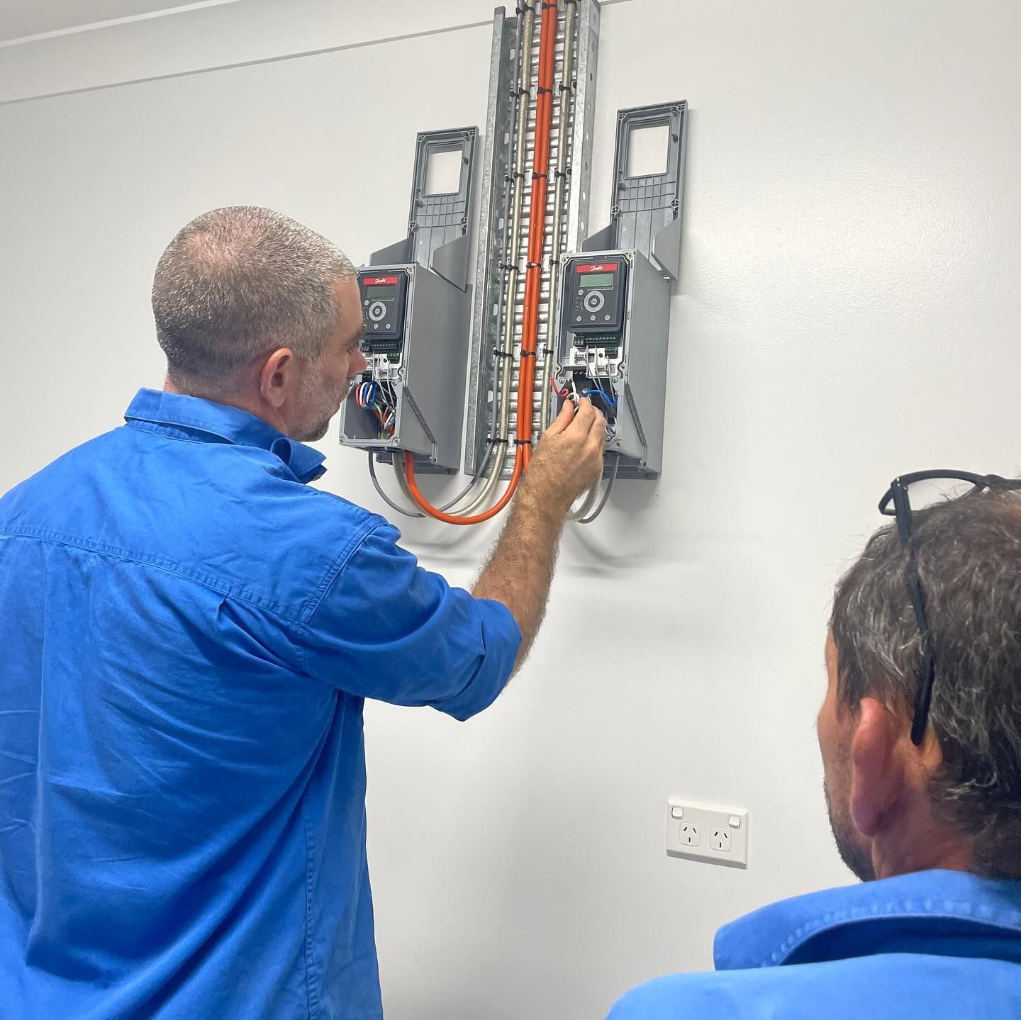 Two men in blue shirts examine electrical equipment mounted on a white wall. One adjusts a dial.— Lahtinen Electrical & Refrigeration in Ingham, QLD