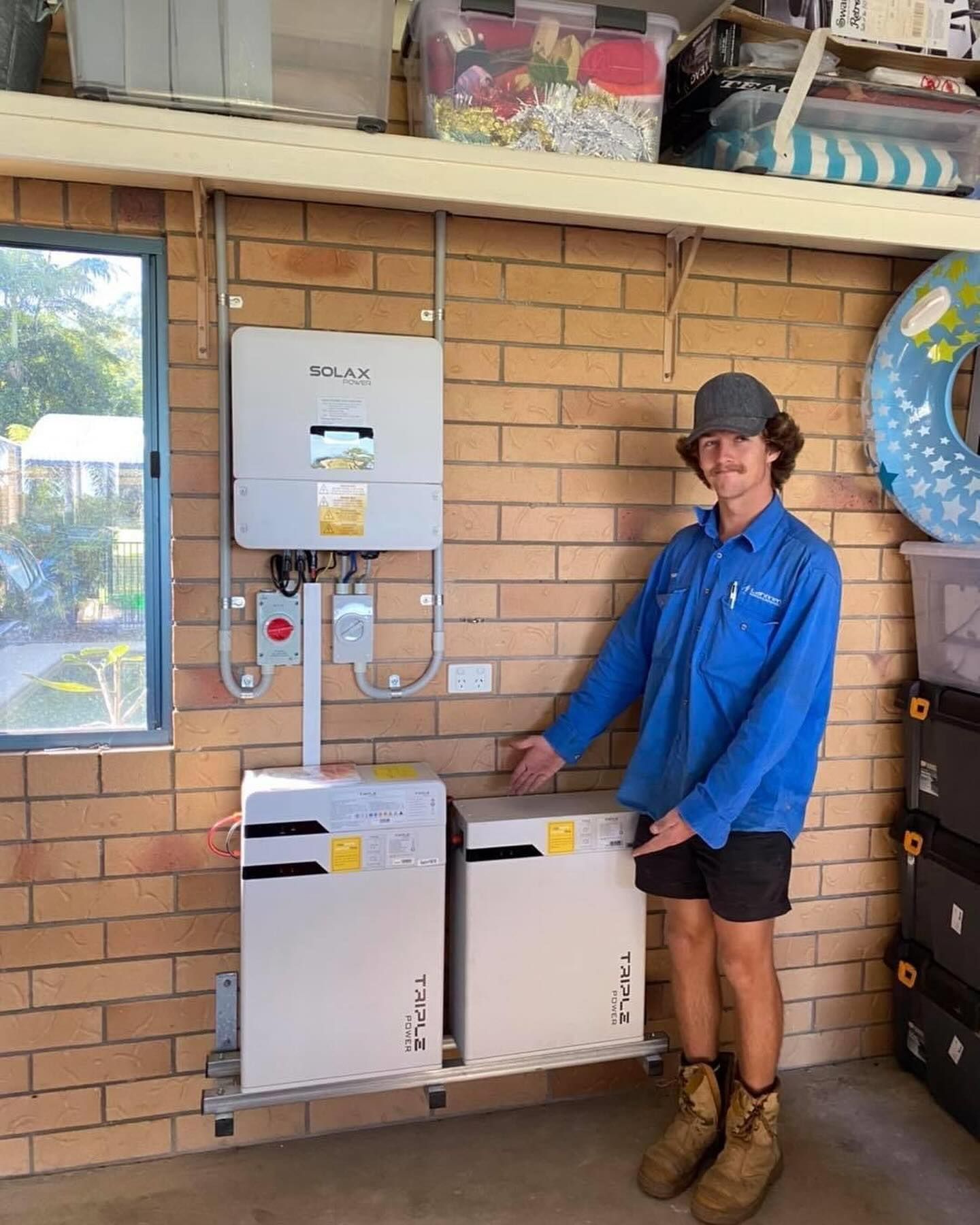Man Points at Two White Solar Battery Units, Next to a Solar Inverter — Lahtinen Electrical & Refrigeration in Ingham, QLD