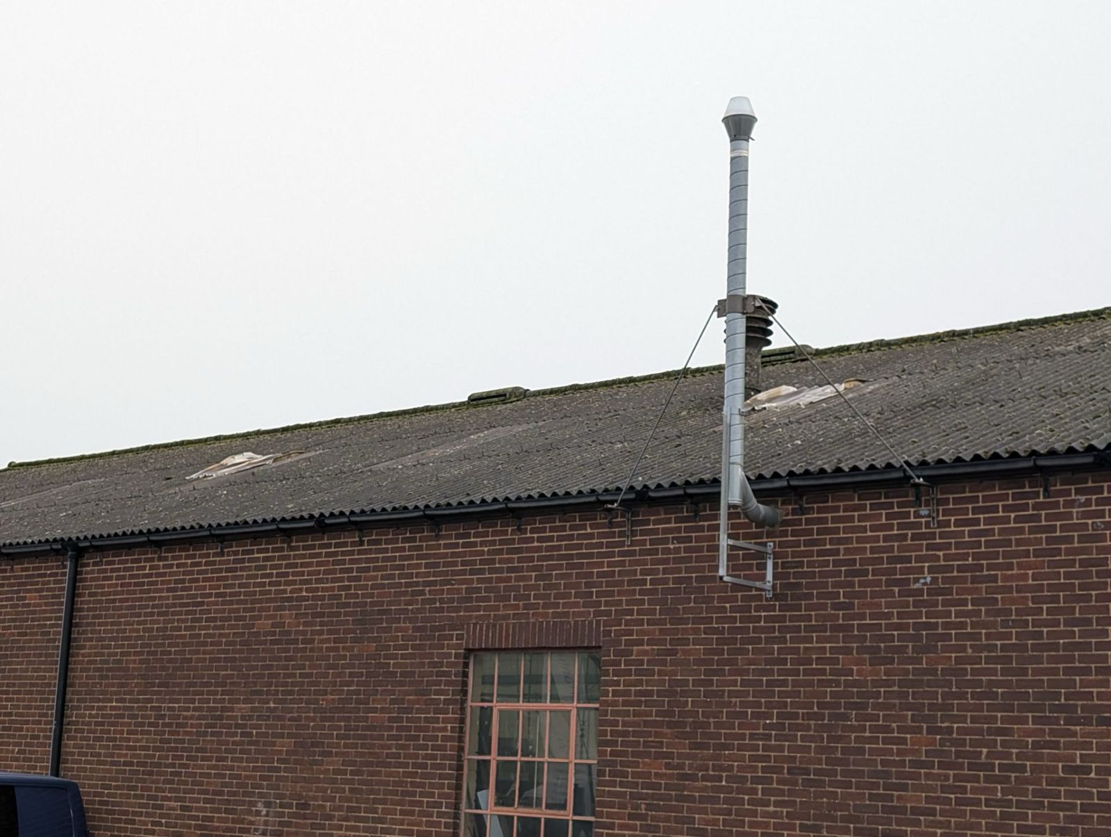 Brick building roof with a tall metal vent pipe and ladder against a cloudy sky