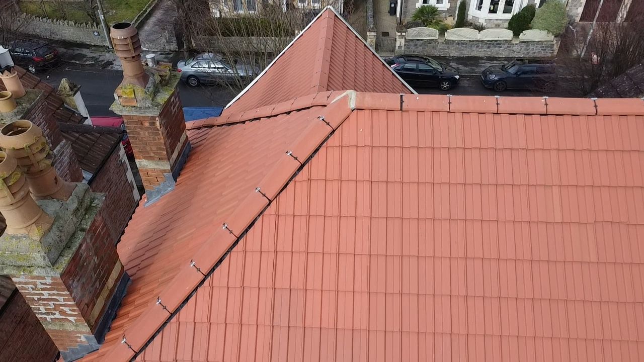 Red-tiled house roof with a diagonal ridge and chimneys beside a courtyard wall