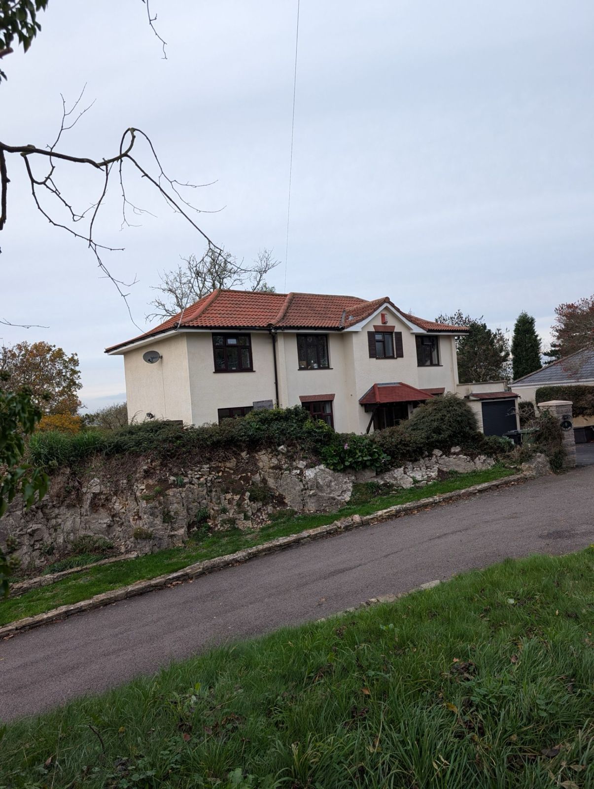 White house with red-tiled roof beside a stone wall and road on an overcast day