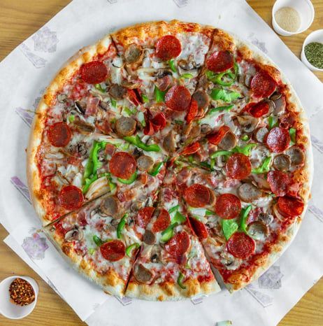 A pepperoni, sausage, and green pepper pizza on paper, with small side containers of seasoning on a wooden surface.