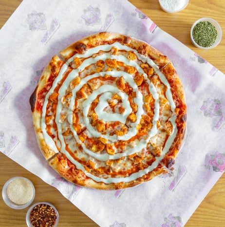 A buffalo chicken pizza drizzled with a white sauce spiral on patterned parchment paper, with spice cups to the side.