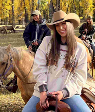 Woman in a cowboy hat riding a horse outdoors with other riders and autumn trees behind her