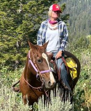 Person riding a brown horse on a forest trail, wearing a red cap and plaid shirt