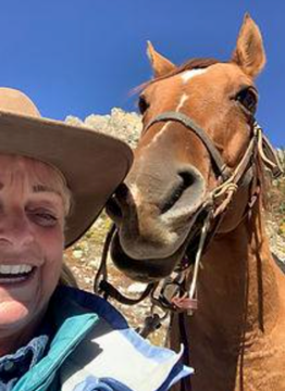 Smiling person in a tan hat beside a chestnut horse under a bright blue sky