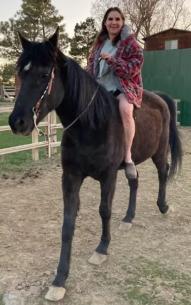 Person riding a black horse outdoors in a fenced area, smiling and wearing a red patterned blanket.