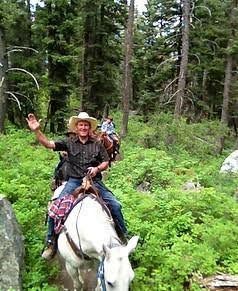 Man riding a white horse on a forest trail, waving, with a small child on the saddle behind him.