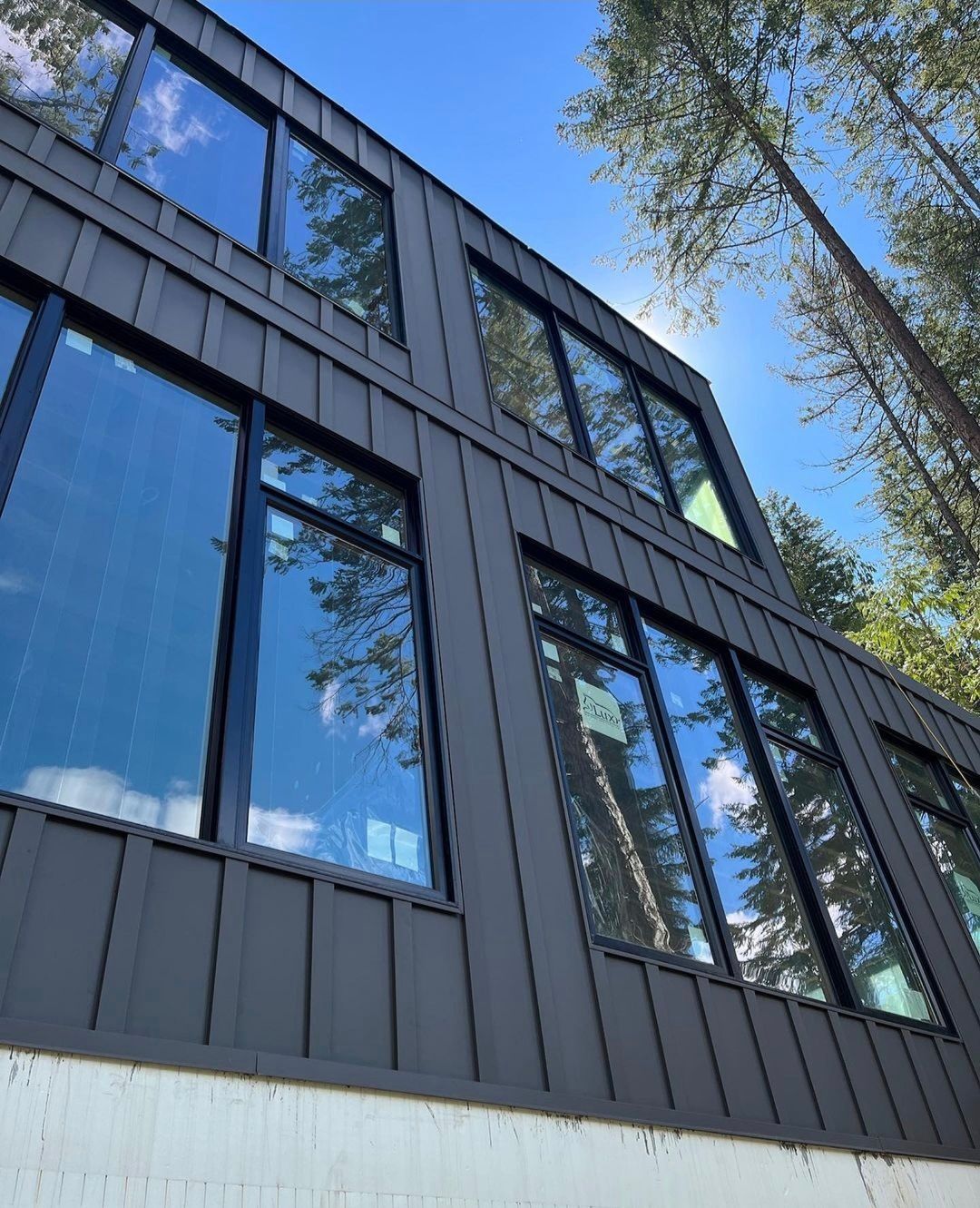 Modern two-story building with dark siding and large windows, reflecting the sky and surrounding trees.