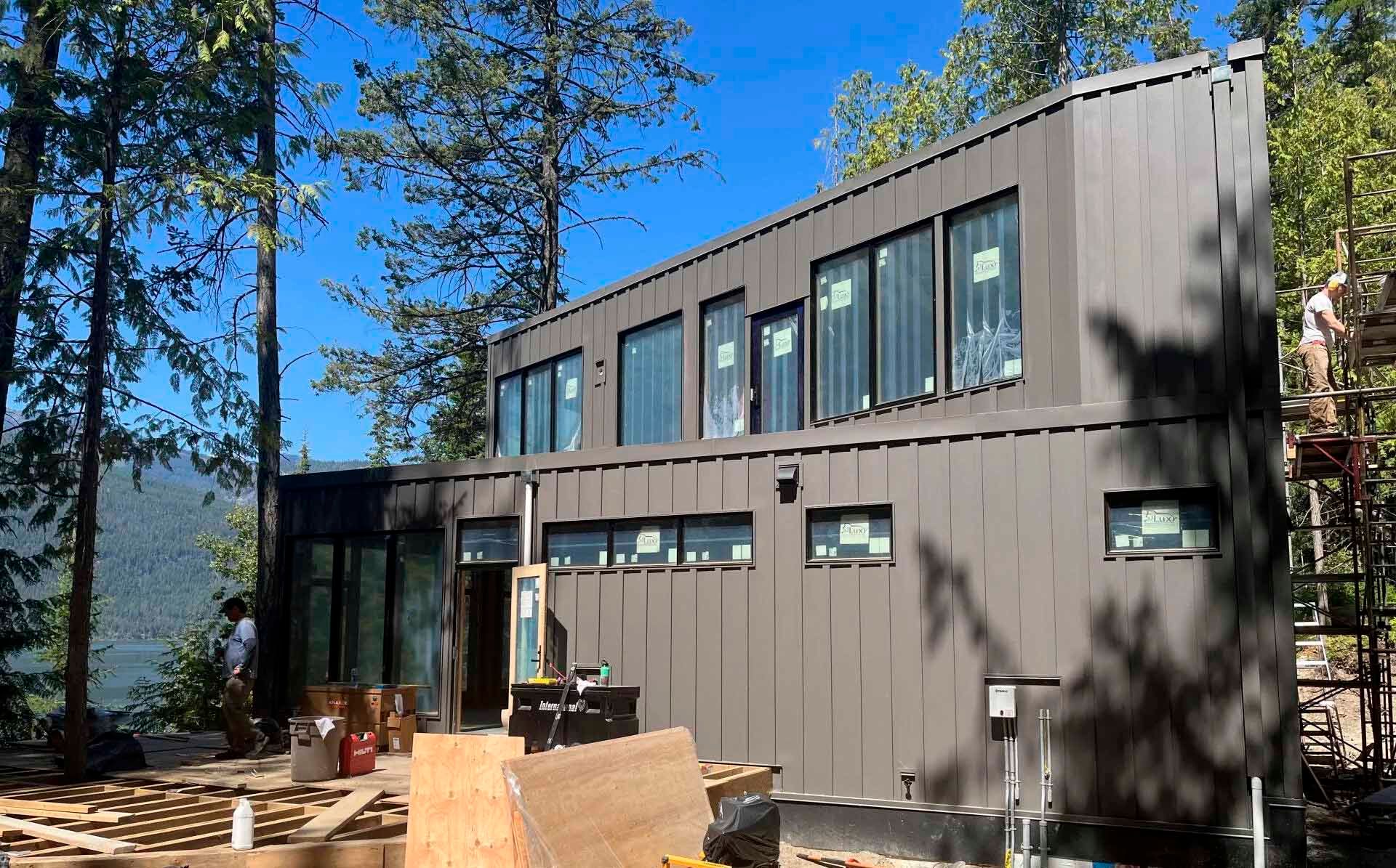 Modern, two-story house under construction with dark gray siding and large windows, surrounded by trees.