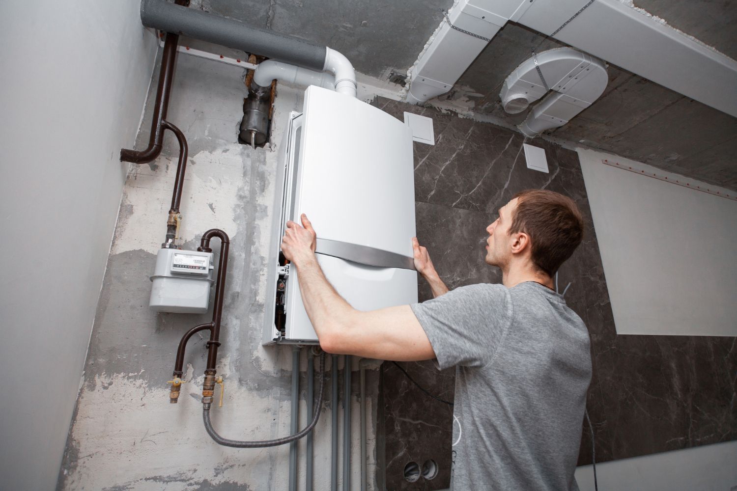 Man installing a white wall-mounted boiler in a room with exposed pipes and unfinished walls.