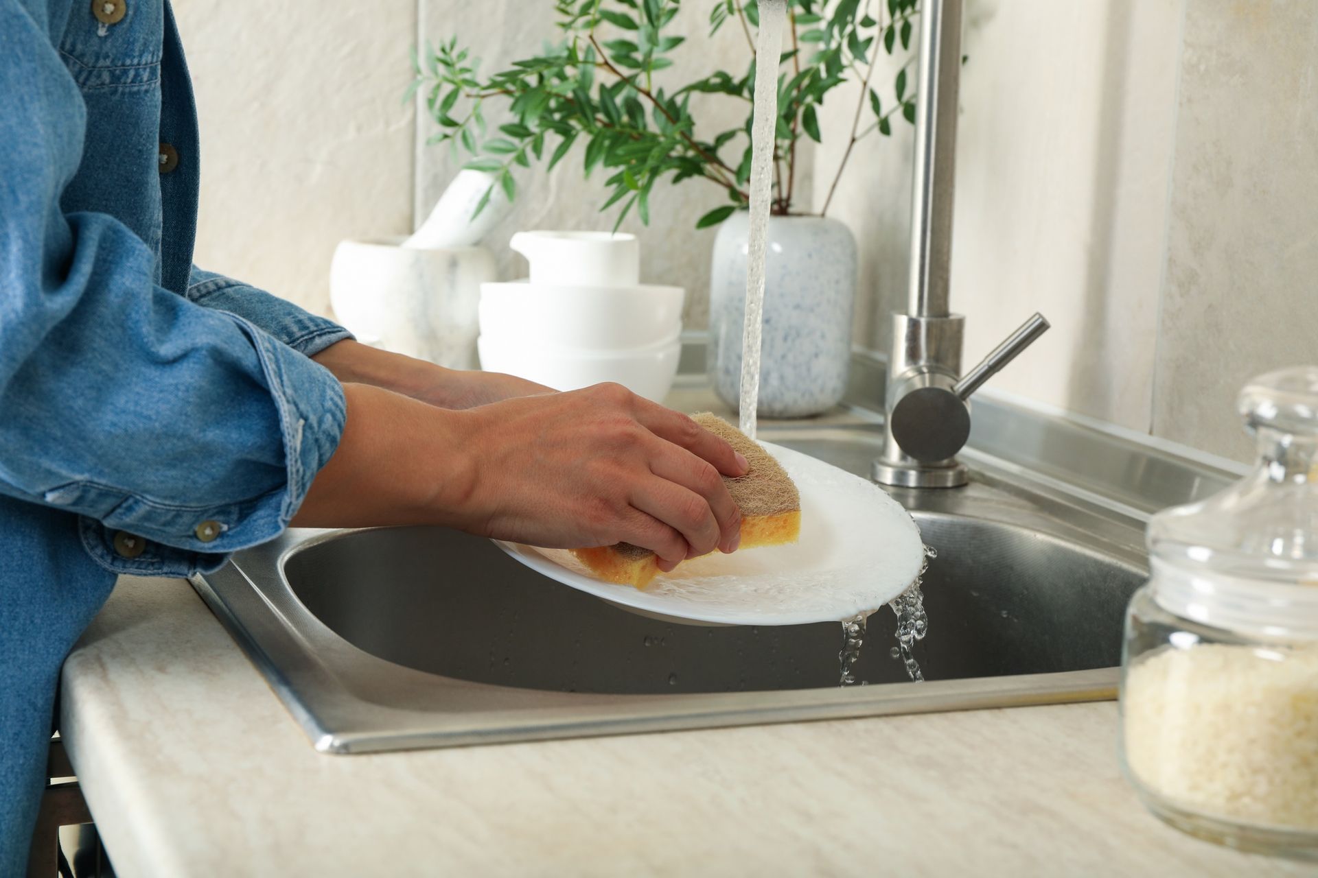 Person washing a white plate in a stainless steel sink, water running from the faucet.