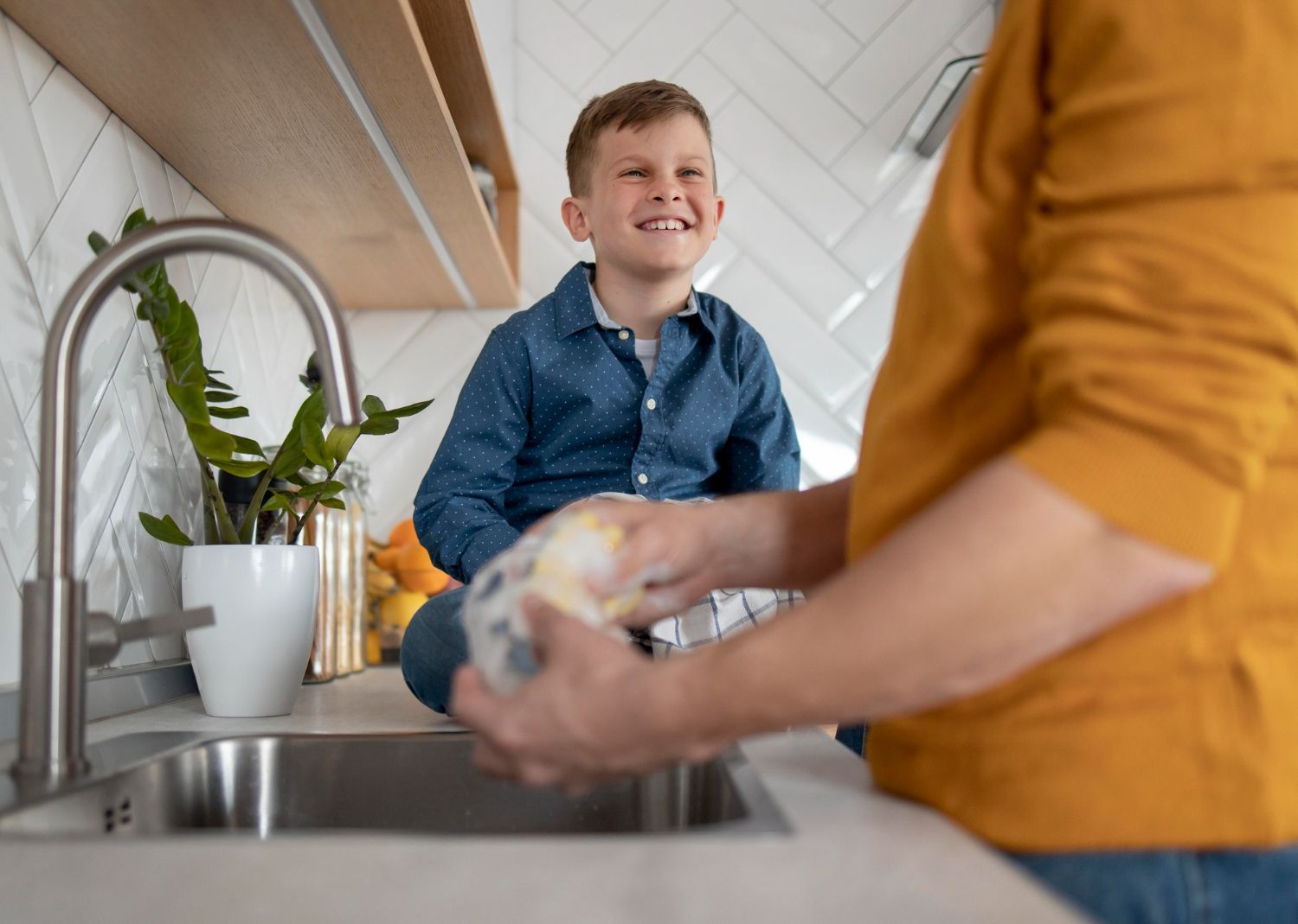 Boy sits on countertop, smiling, as person washes something in kitchen sink.